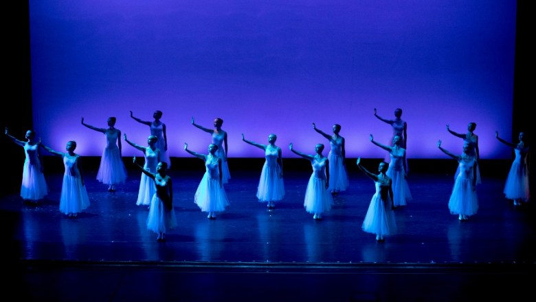 A group of fifteen ballet dancers in long white tutus perform on stage, set against a blue gradient background. Their arms are raised in various poses.