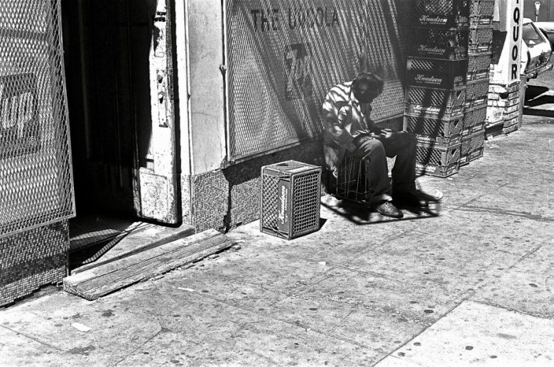 A person sits on a crate outside a liquor store in bright sunlight, looking down, with stacked boxes and a shadow cast on the sidewalk.