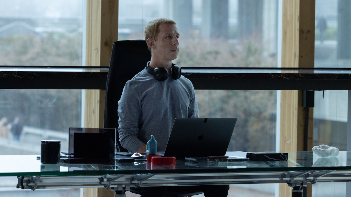 a white man in a gray sweatshirt sits very upright at a desk with large headphones around neck