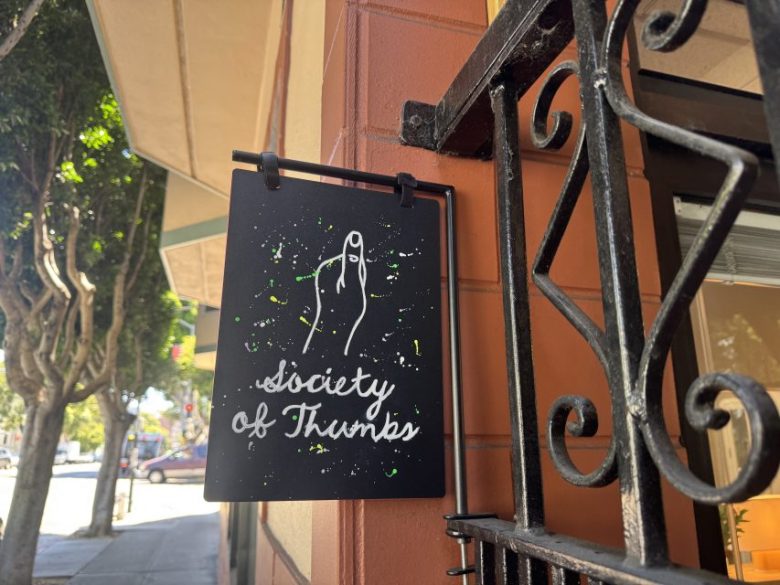 A black sign with a white drawing of a hand showing a thumbs up and the text “society of thumbs” hangs on a metal bracket outside a building.