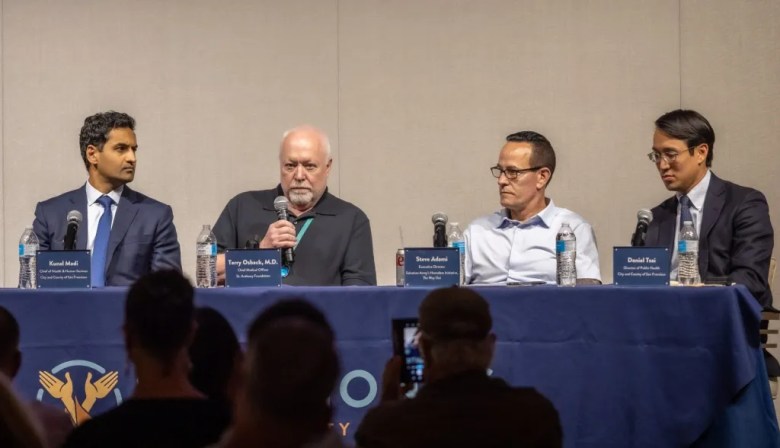 Six people sit on stage before an audience at a public forum.