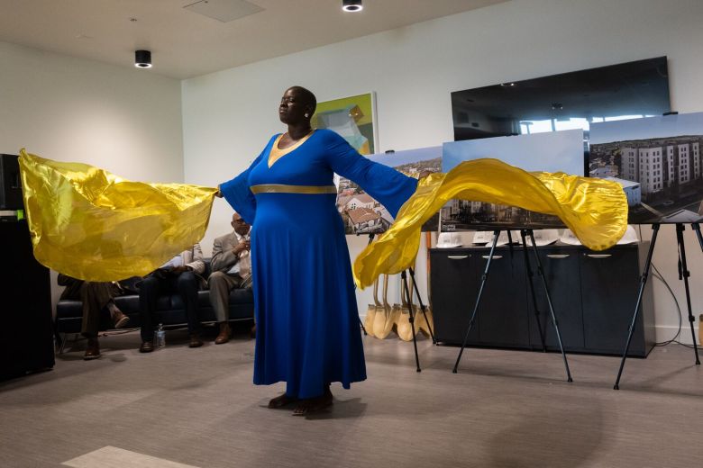 A woman dressed in blue holds billowing sheets of gold fabric as she dances in front of renderings of a new apartment building