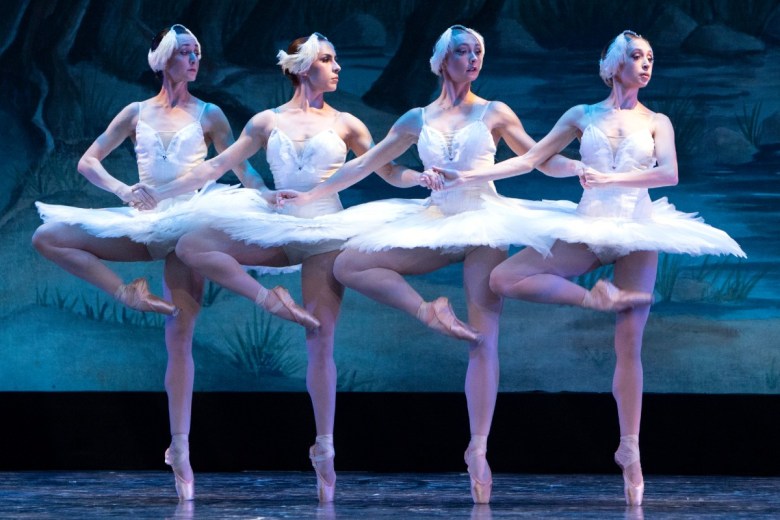 Four female ballet dancers in white tutus perform together on stage against a blue and green backdrop, showcasing elegant poses and leg extensions.