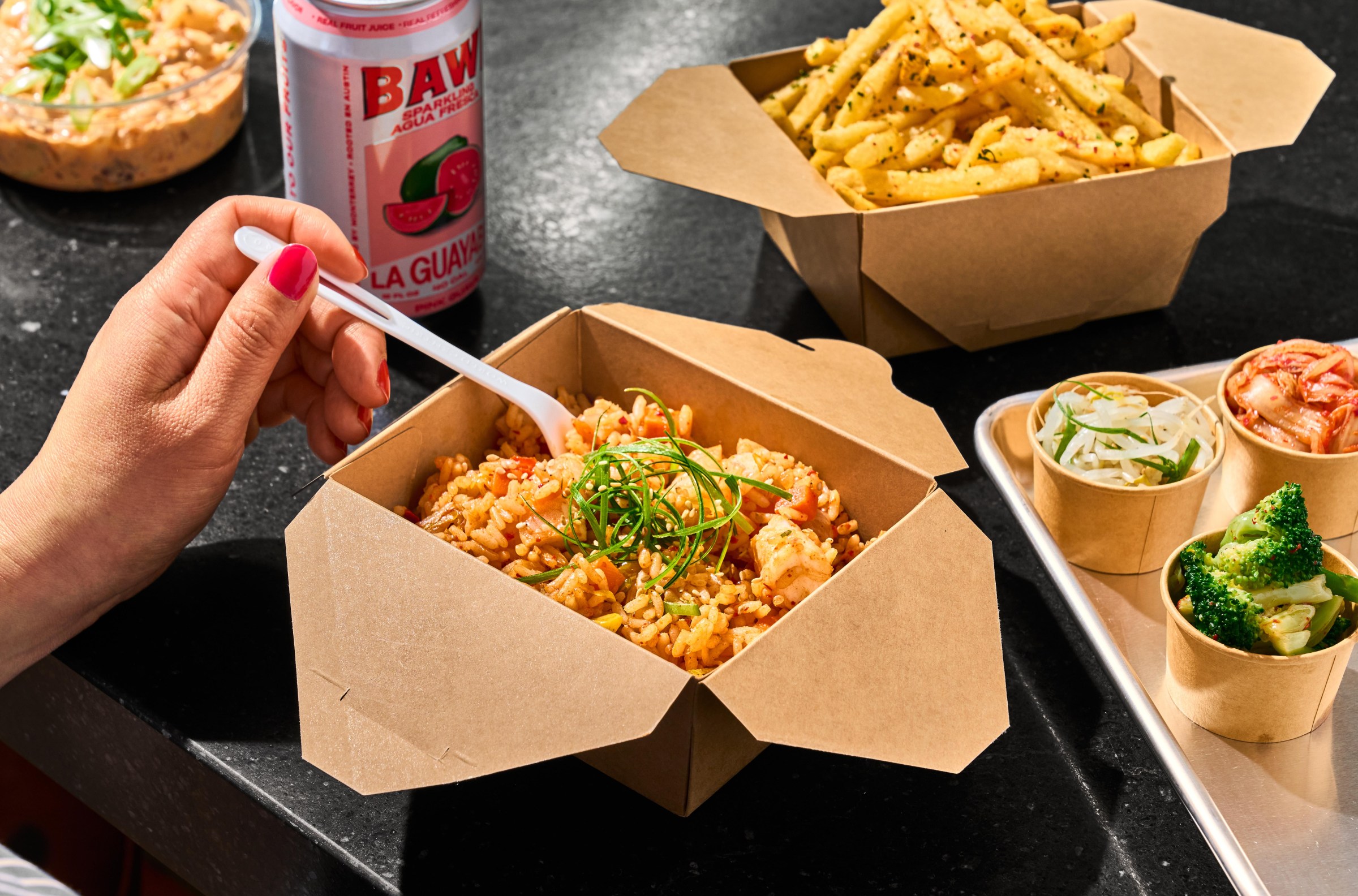 A hand holds a fork inside a box of kimchi rice, with fries and banchan alongside at the Mess Hall in San Francisco.
