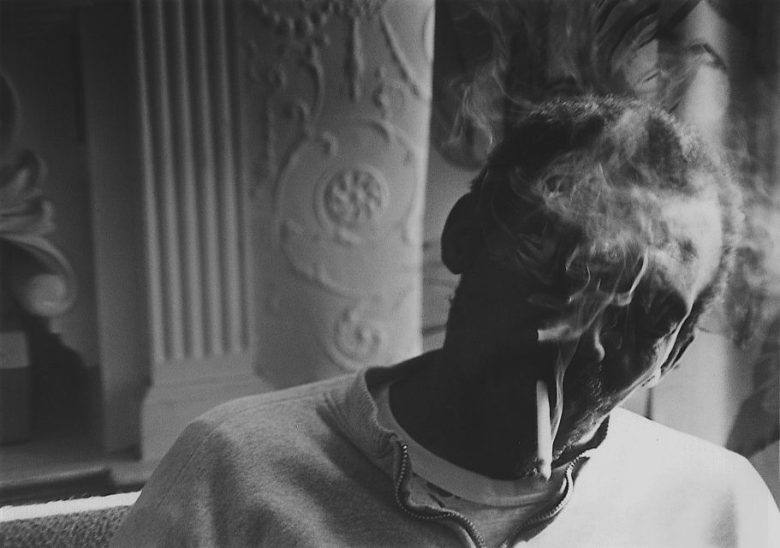 A man sits indoors with smoke partially obscuring his face. Ornate architectural details are visible in the background.