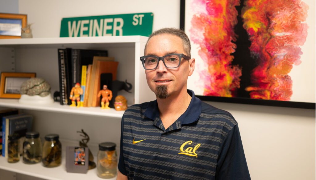 man with soul patch, glasses and Cal shirt standing in front of colorful painting