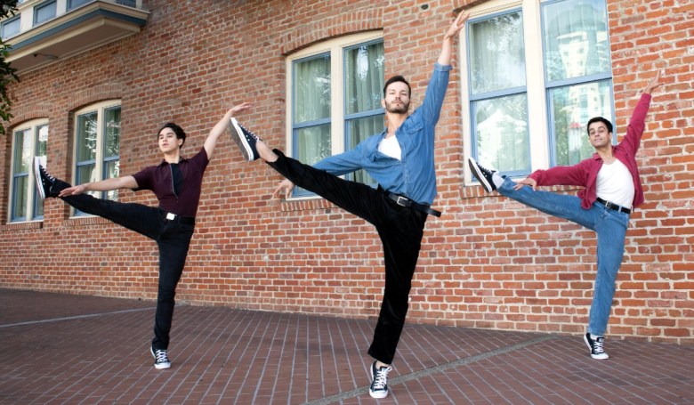Three men in colorful outfits perform a synchronized dance pose in front of a brick wall, with legs extended and arms raised.