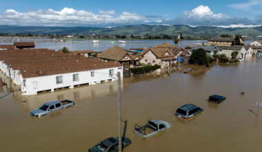 An aerial shot of houses, streets and cars completely submerged in brown flood water.