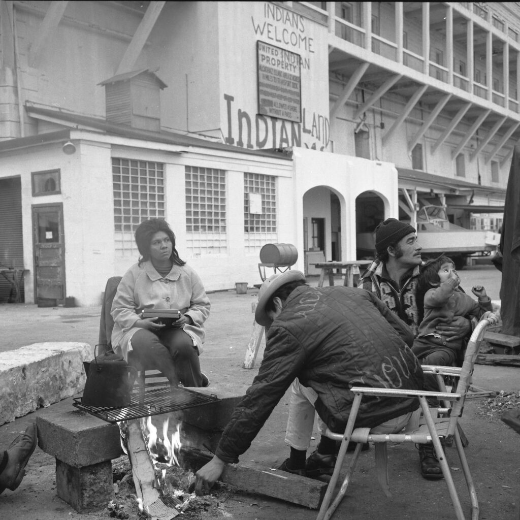 A black and white photograph showing a group of Native American occupiers keeping warm around a small campfire in a paved dock area on Alcatraz Island. One person tends the fire, while a woman sits nearby holding a small box, and a man sits holding a young child. A coffee pot rests on a grill over the flames. Behind them, a white building features painted graffiti reading "INDIANS WELCOME," "UNITED INDIAN PROPERTY," and "INDIAN LAND," altering an official sign.