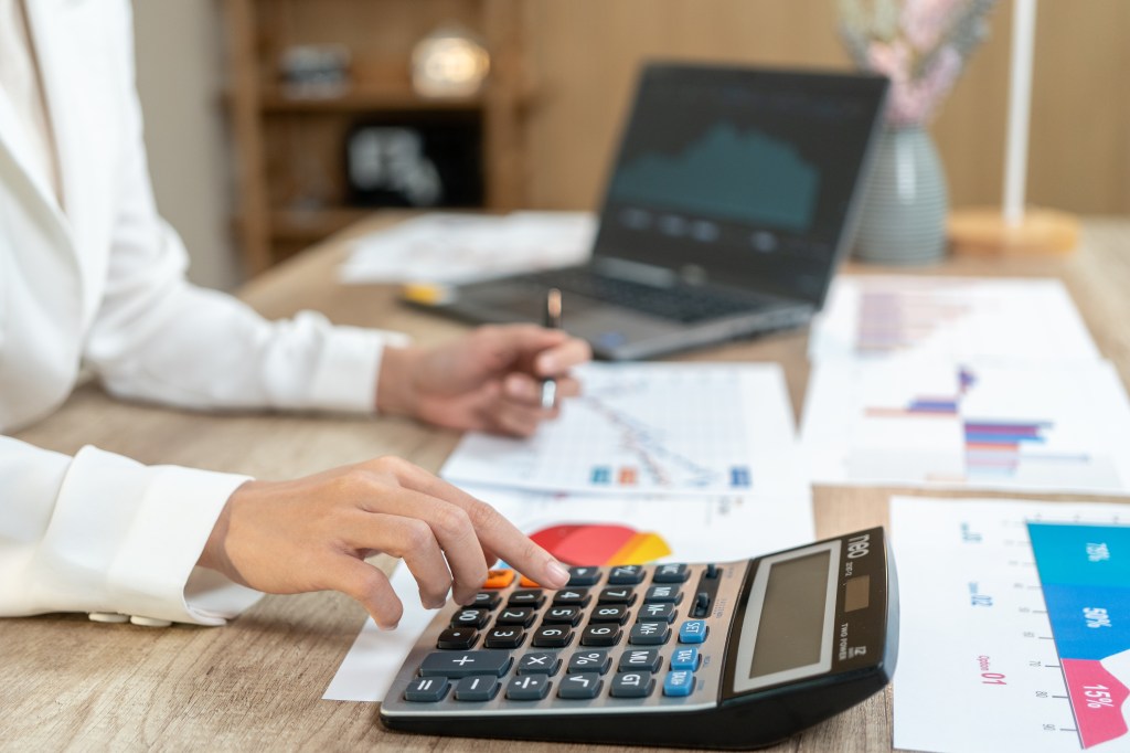 A person using a calculator, with financial documents and a laptop in the background.