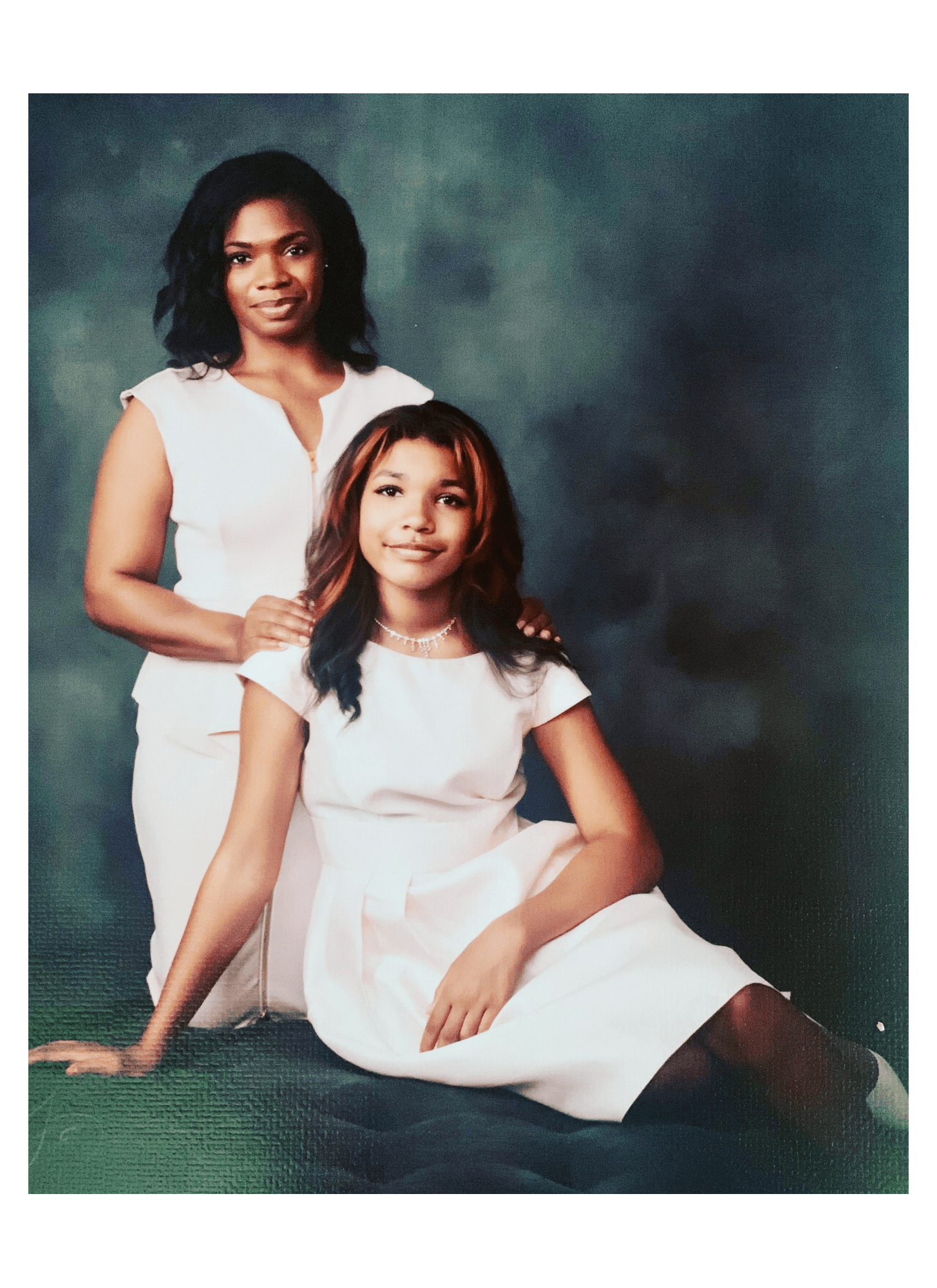 Two women, one sitting and one standing, pose against a dark background, both wearing white dresses.