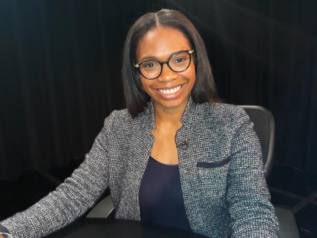 Smiling woman with glasses and shoulder-length hair, wearing a gray blazer and navy top, sitting at a desk.