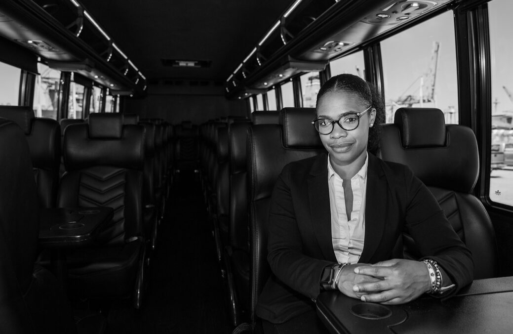 Woman in glasses and formal attire sitting at a table inside a bus, with windows and empty seats around her.