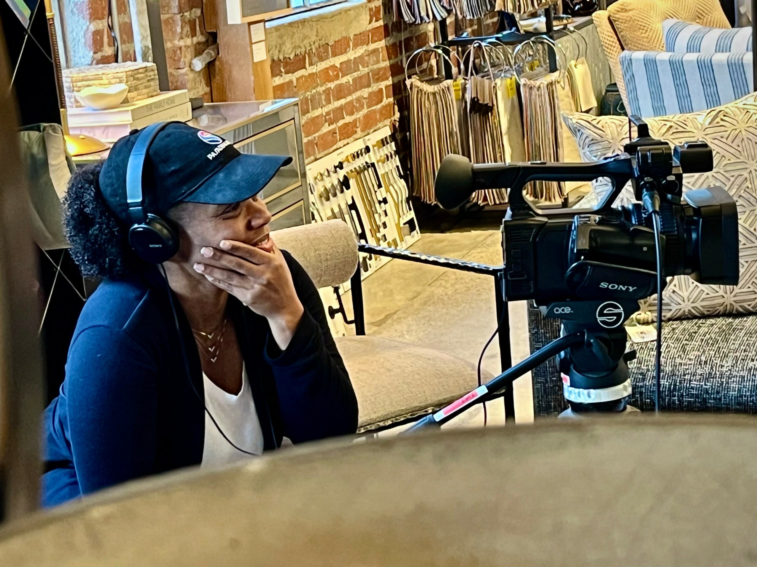 Woman wearing headphones and a cap, sitting in a room with furniture and brick wall, speaking into a camera microphone.