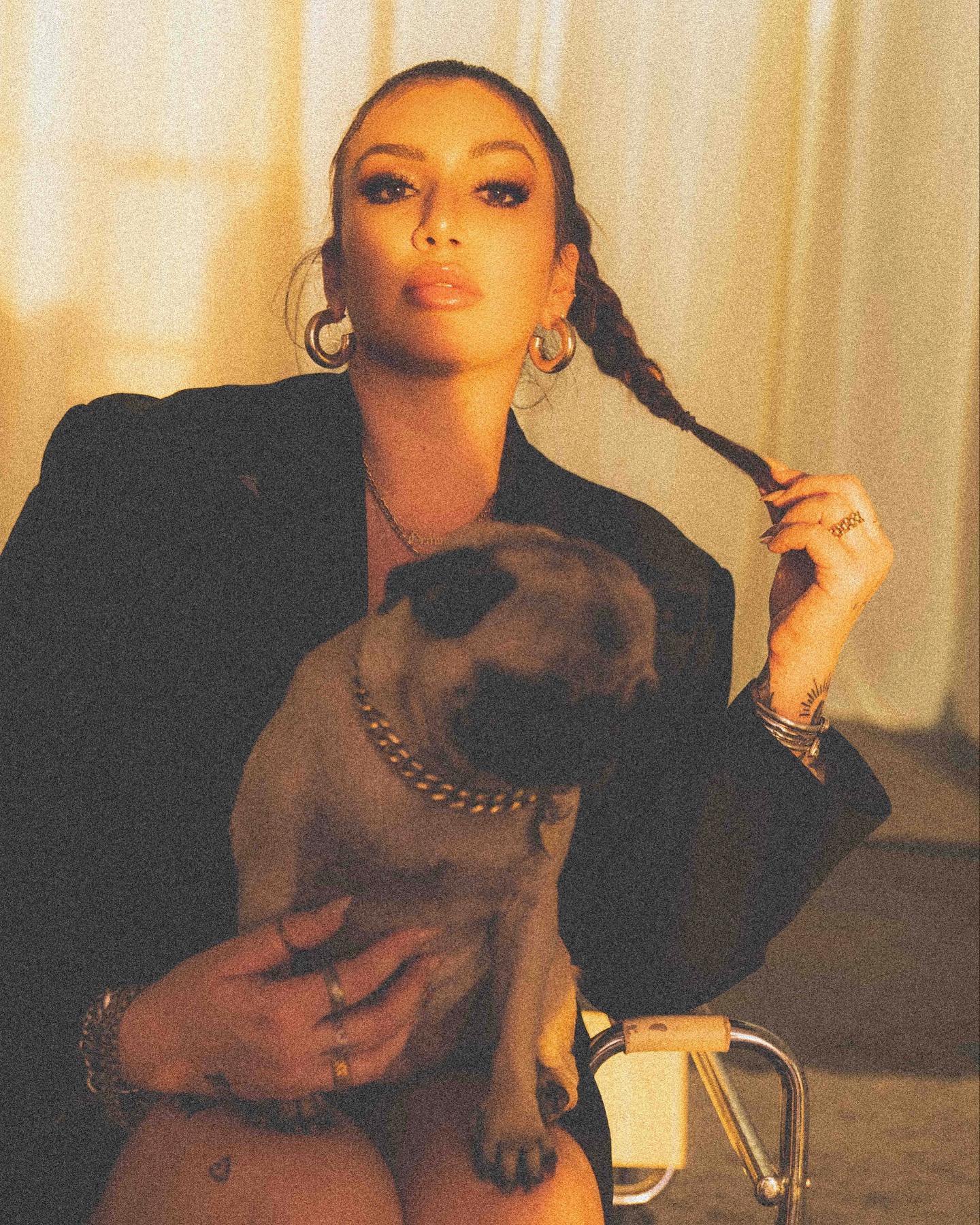 Woman with braided hair holding a dog, wearing a black blazer, in front of a curtain background.