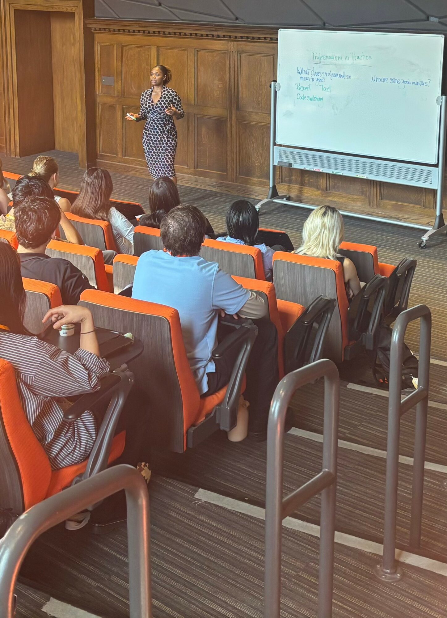 Woman standing and speaking to an audience in a lecture hall with a whiteboard, audience seated in orange and black chairs.