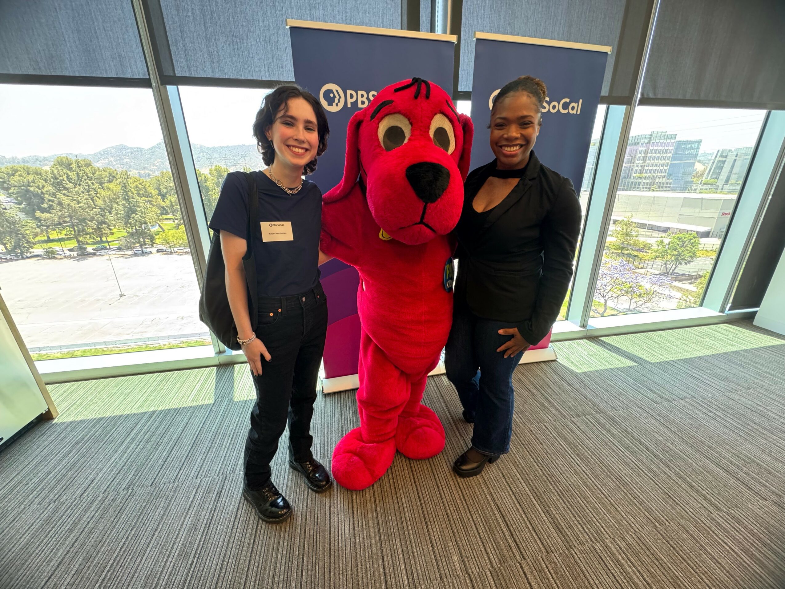 Two women and a person in a red dog mascot costume stand indoors near large windows, smiling at the camera.