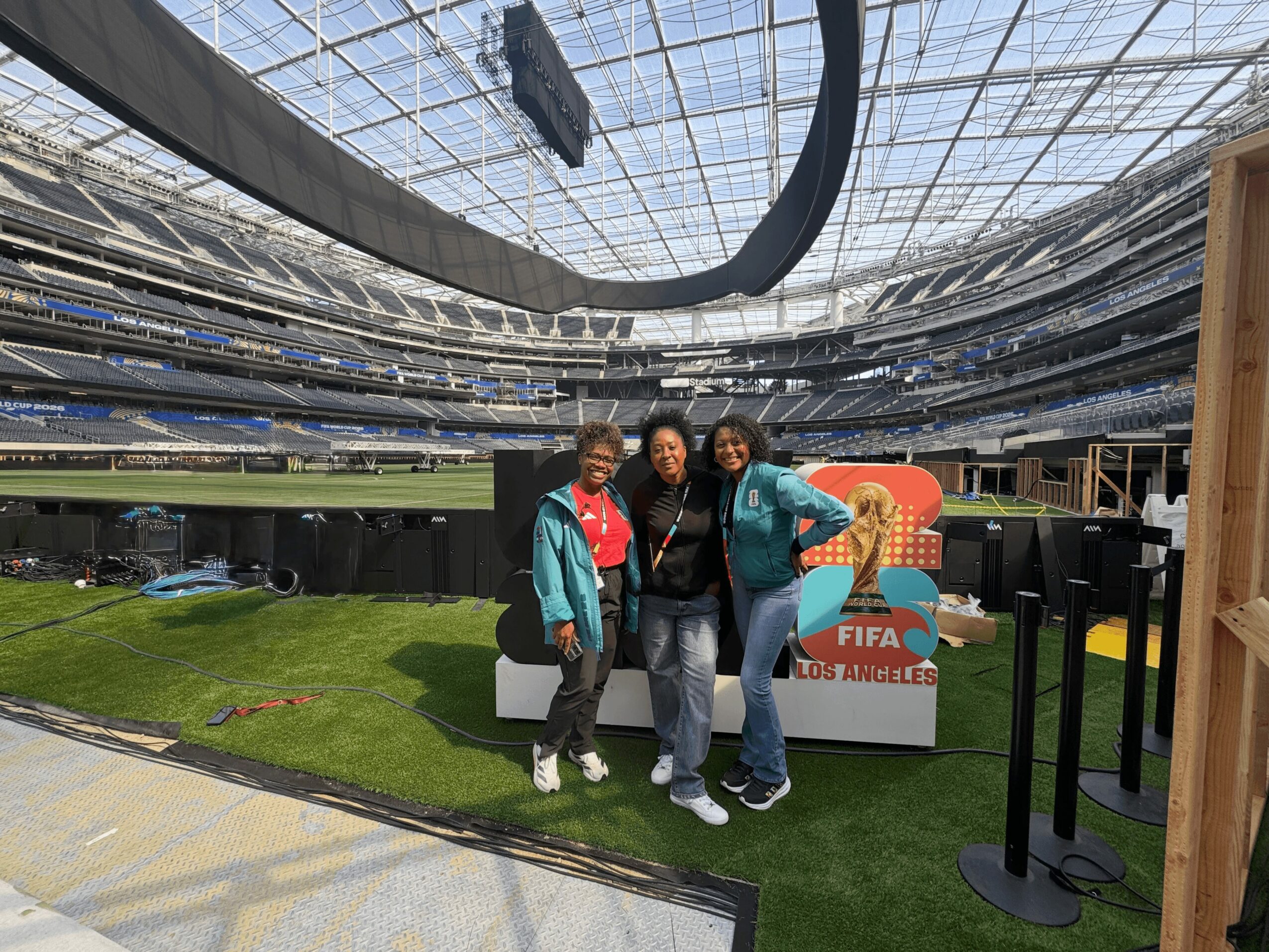 Three people standing on artificial grass in a stadium, with a FIFA Los Angeles sign behind them, under a glass roof.