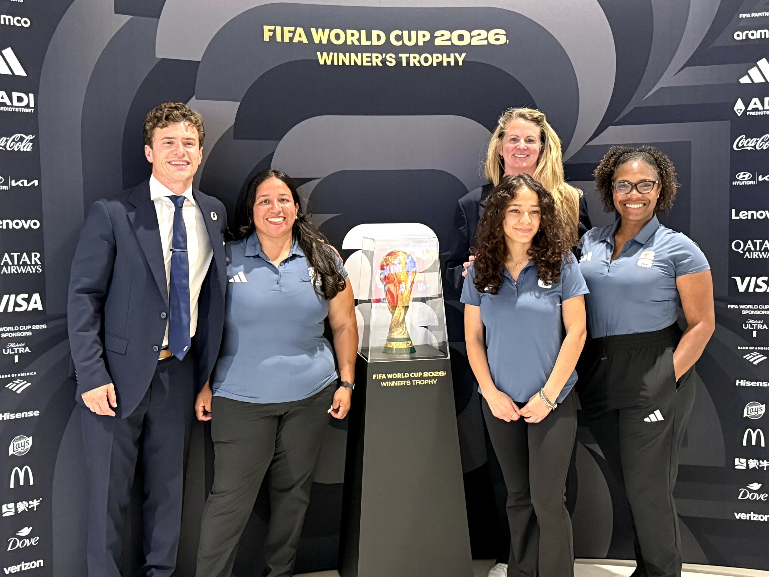 Group of five people standing in front of FIFA World Cup 2026 winners trophy display, smiling, indoors.
