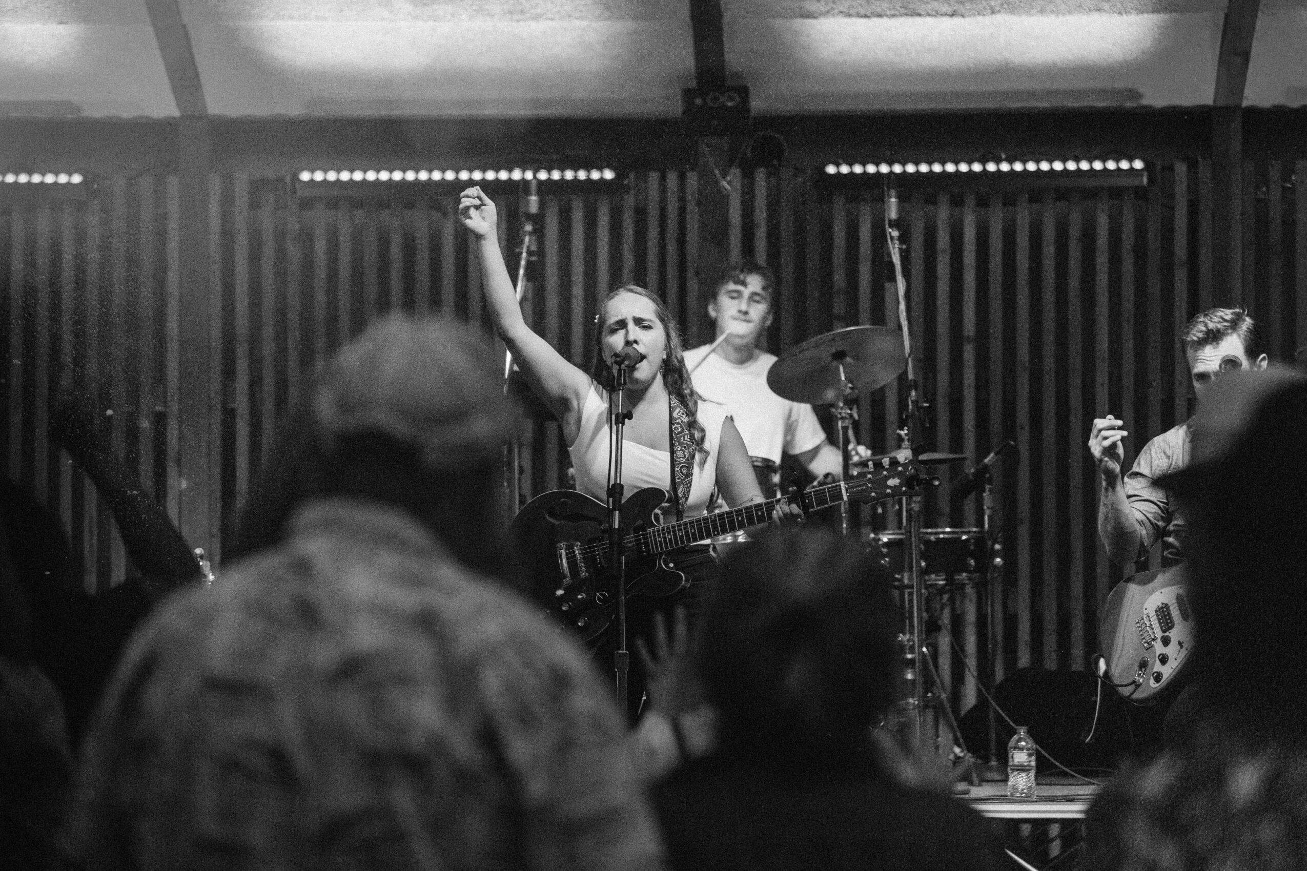 Band performing on stage with audience in foreground, woman singer raising her arm, drummer behind her, and guitarist on the right.