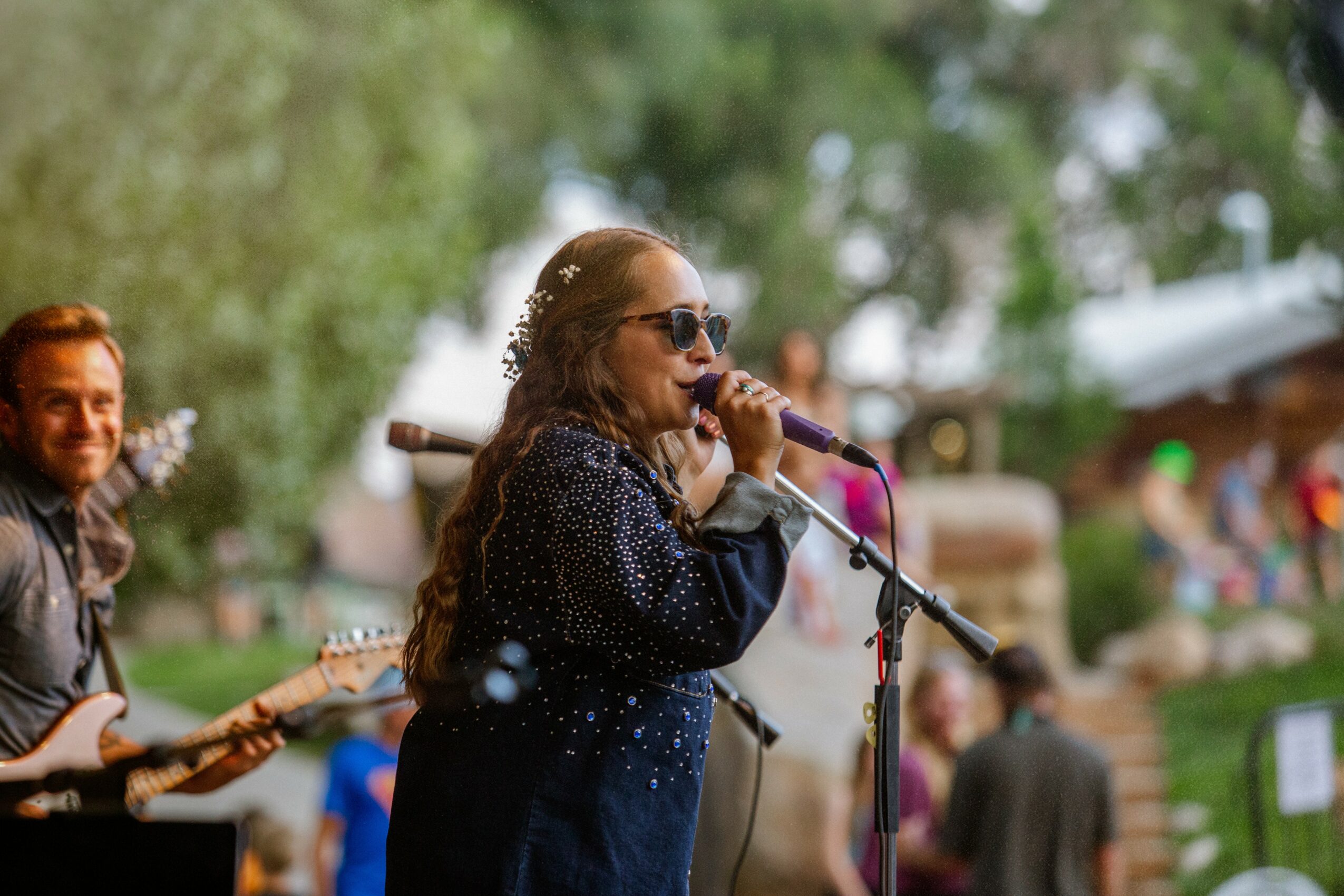 Young girl singing into a microphone outdoors, with a person playing guitar beside her, trees and people in background.