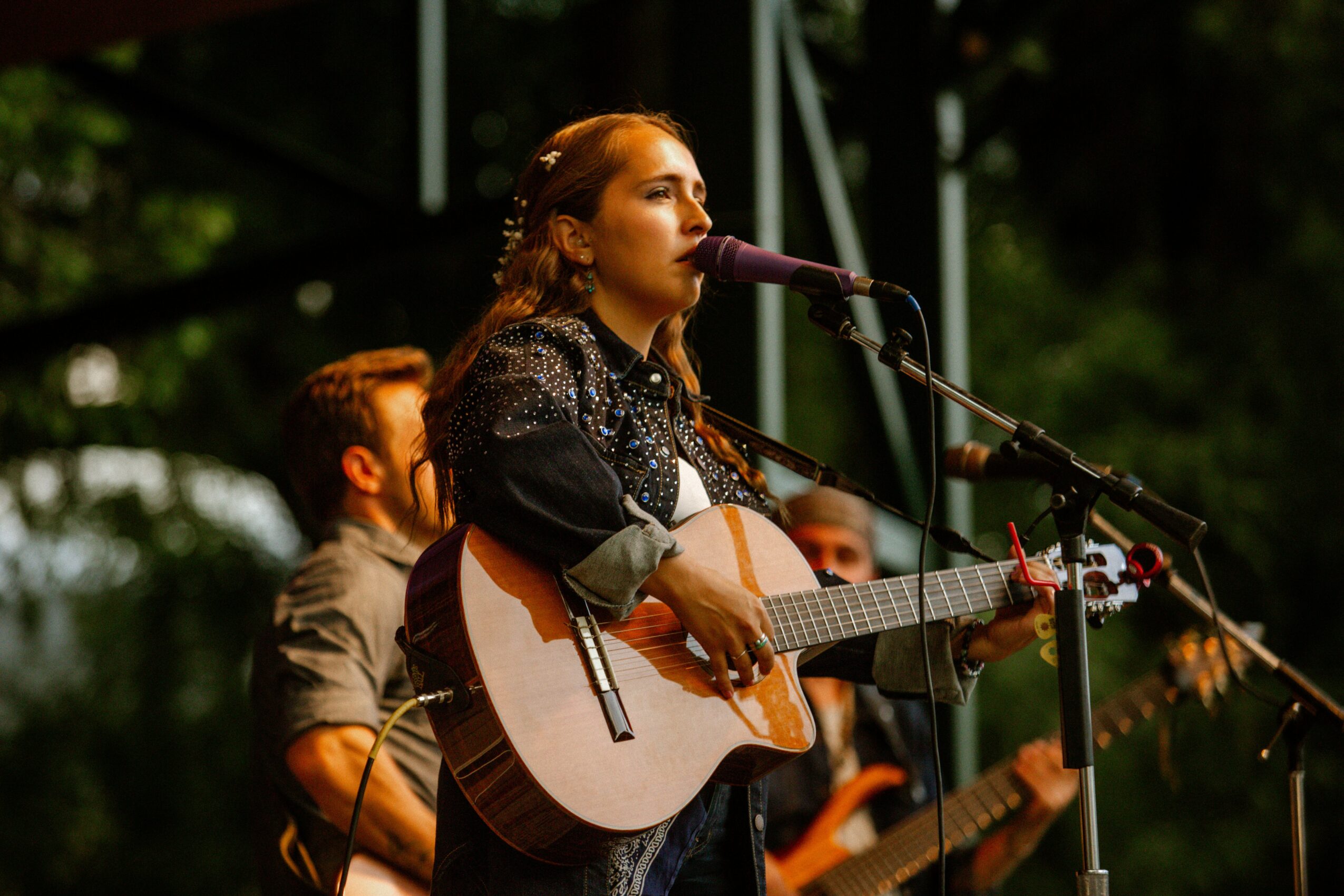 Woman singing and playing guitar on stage with a microphone, with a man and other musicians in the background.