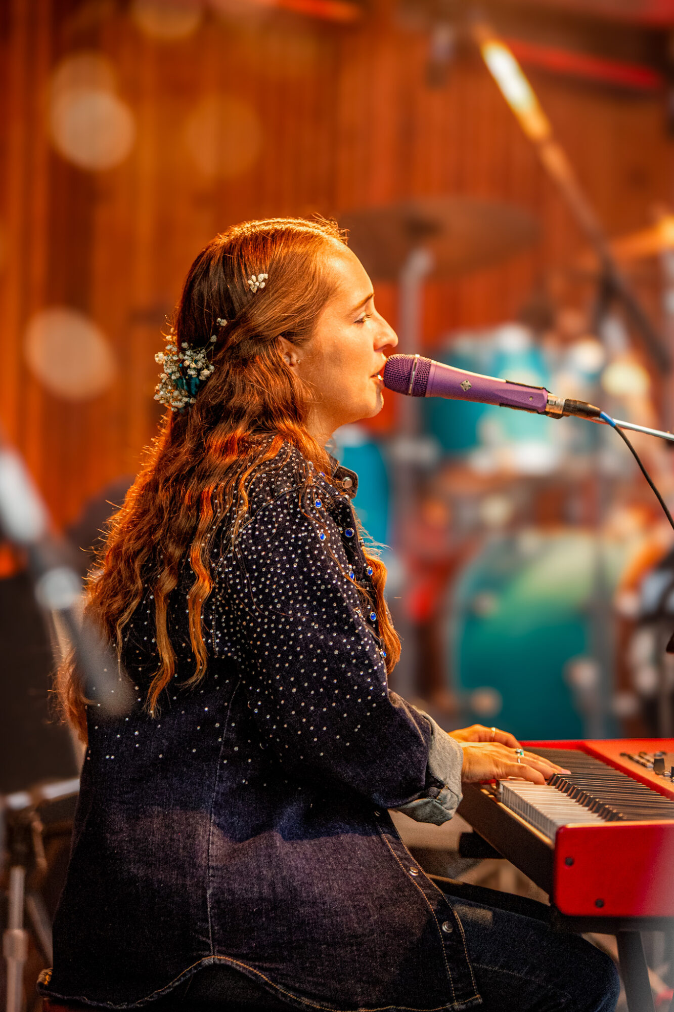 Woman with long hair singing into a microphone while playing a keyboard on stage, warm lighting, blurred background
