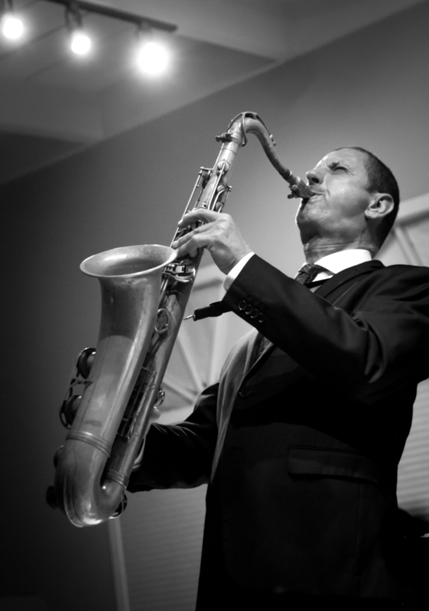 Man in formal suit playing a saxophone on stage, with stage lights overhead, in black and white.