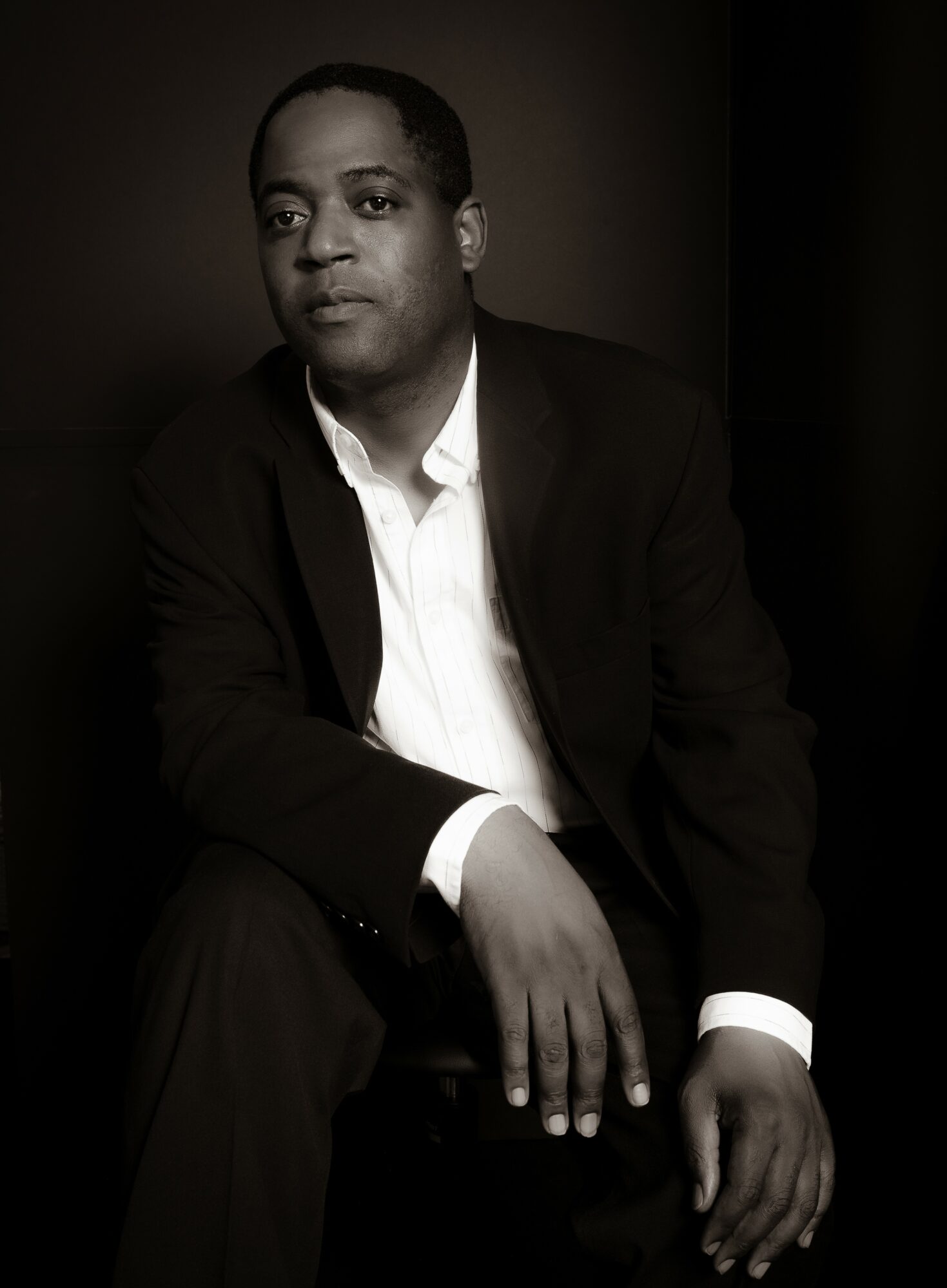 Man in suit sitting with hand on knee, looking at camera, dark background, black and white photo.
