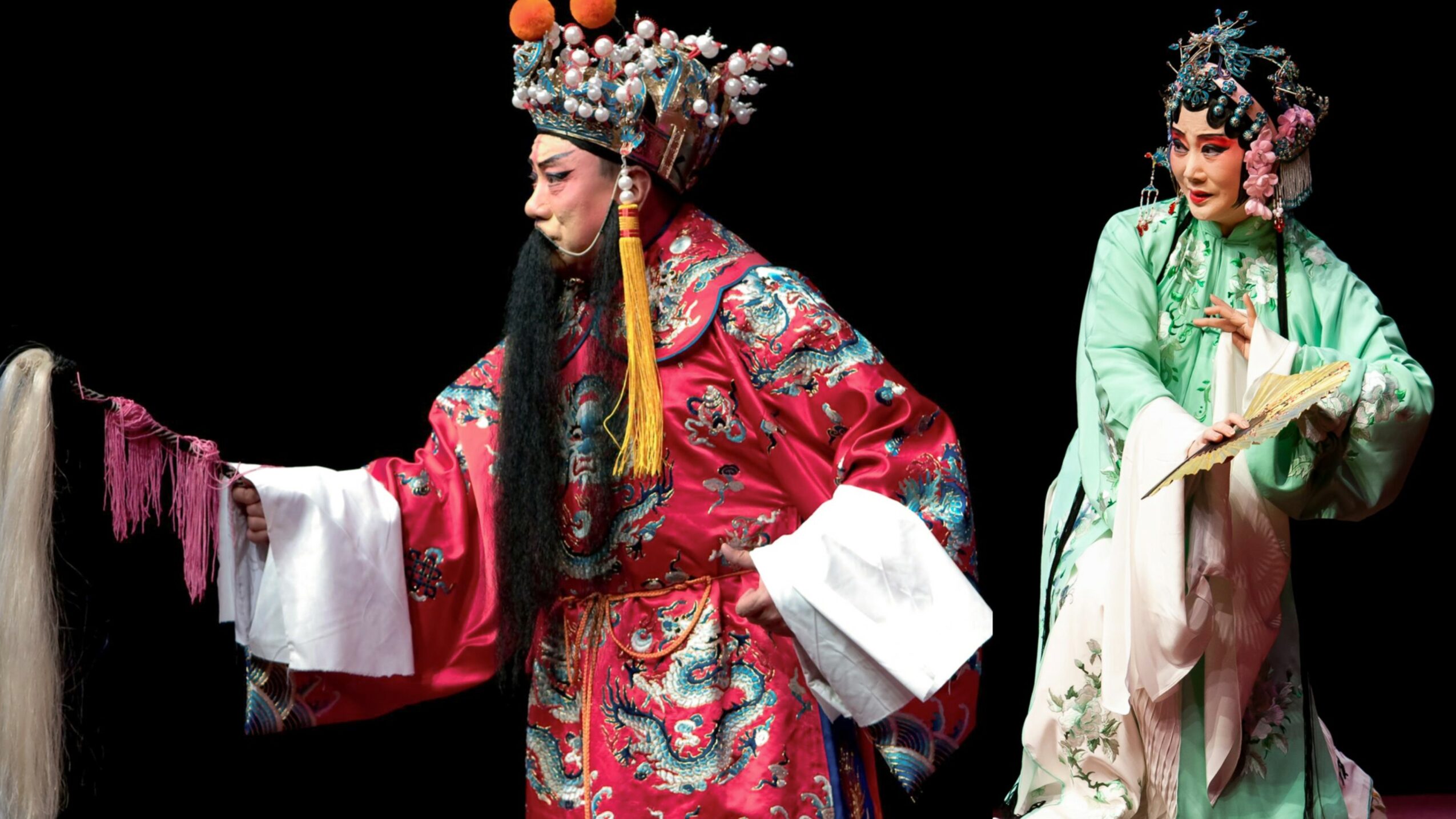 Two performers in traditional costumes and elaborate headdresses on stage, one in red and the other in green, against a black background.