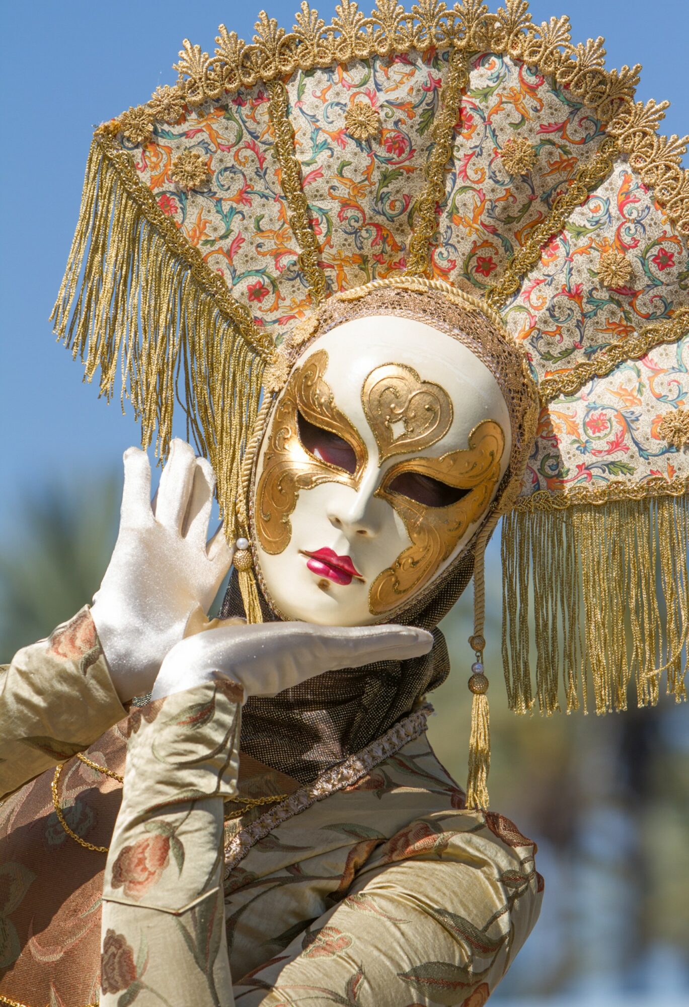 Person wearing an ornate mask and large decorative headpiece with gold and floral patterns, posing outdoors against a blue sky.
