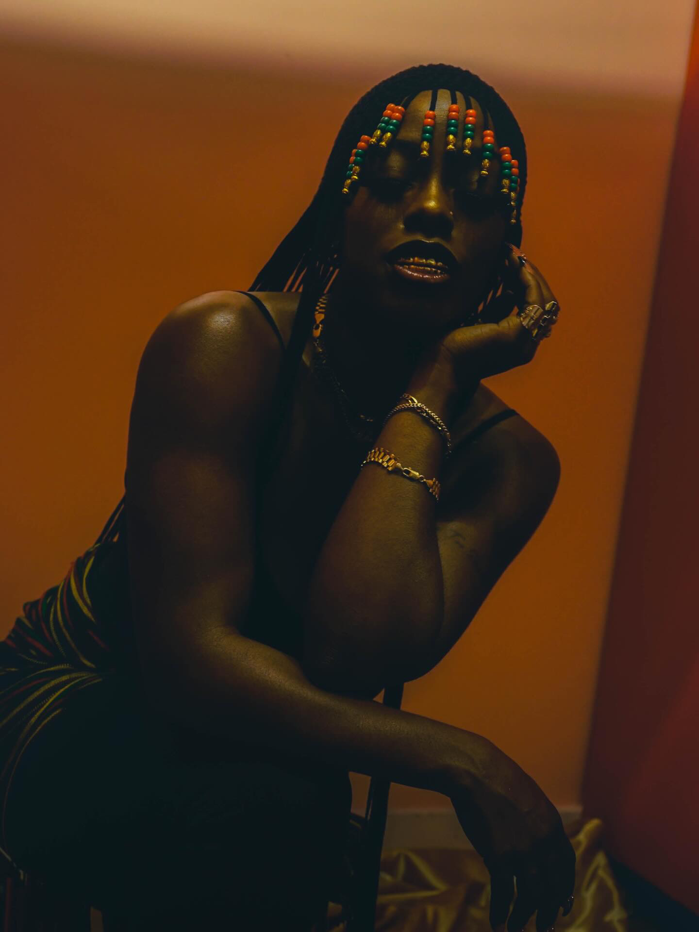 Person with colorful beaded hair accessories, dark makeup, and jewelry, resting chin on hand, in dim lighting.