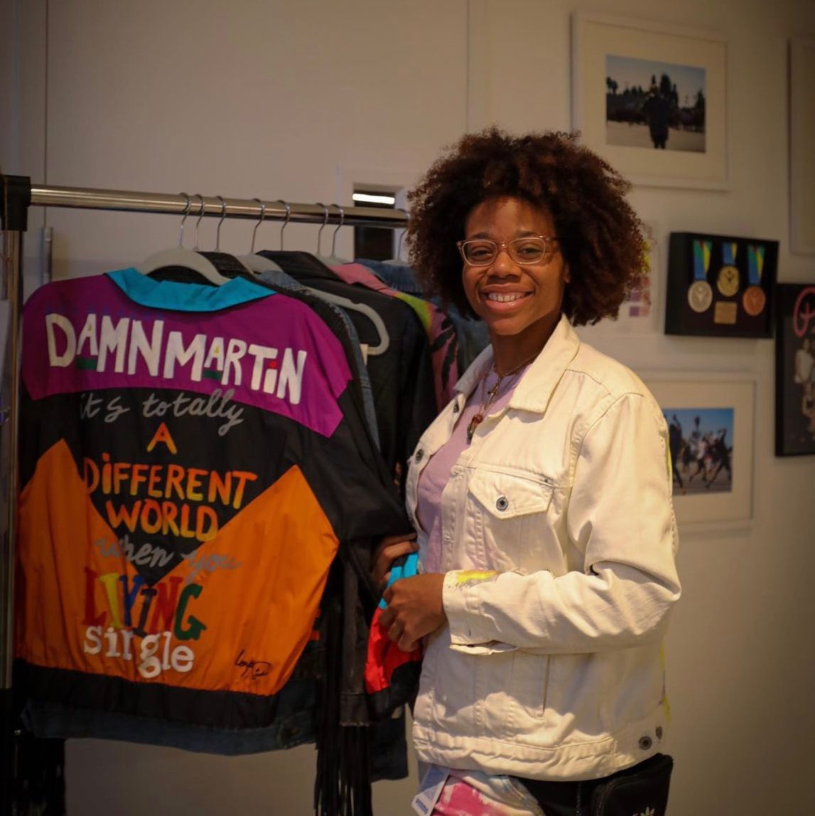 Woman with curly hair and glasses smiling next to a clothing rack with colorful jackets, in an art gallery setting.