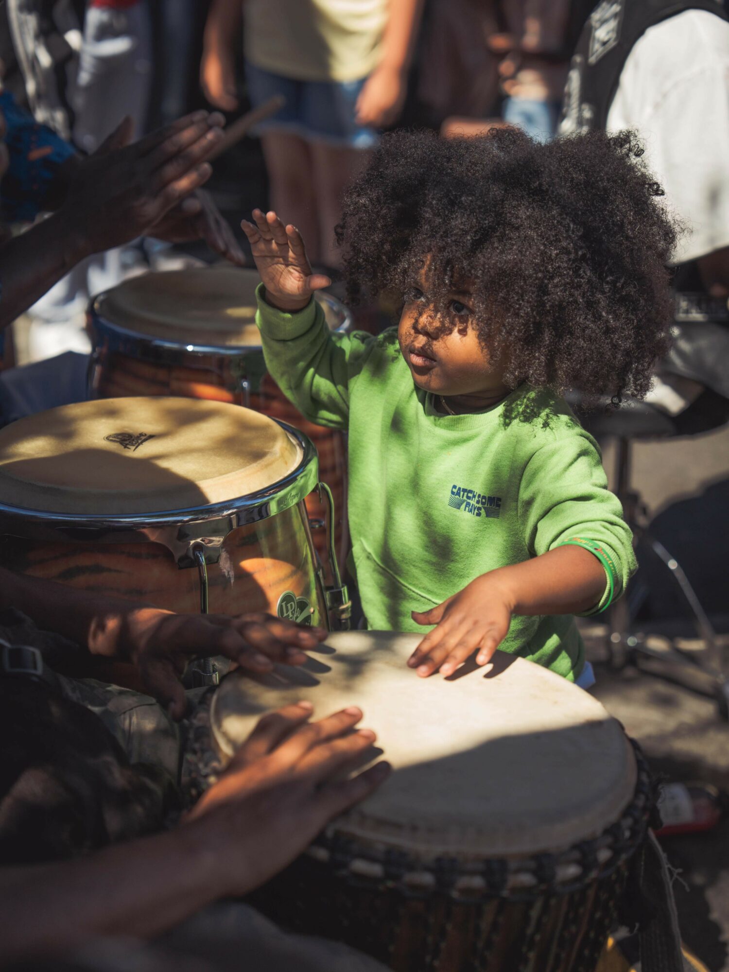 Child with curly hair playing a drum, surrounded by people, outdoors, in sunlight.