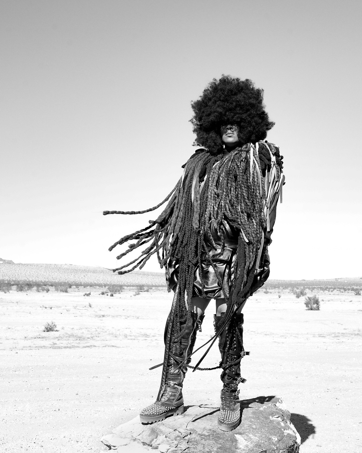 Person with large curly hair and fringed clothing standing on a rock in a desert landscape, clear sky background.