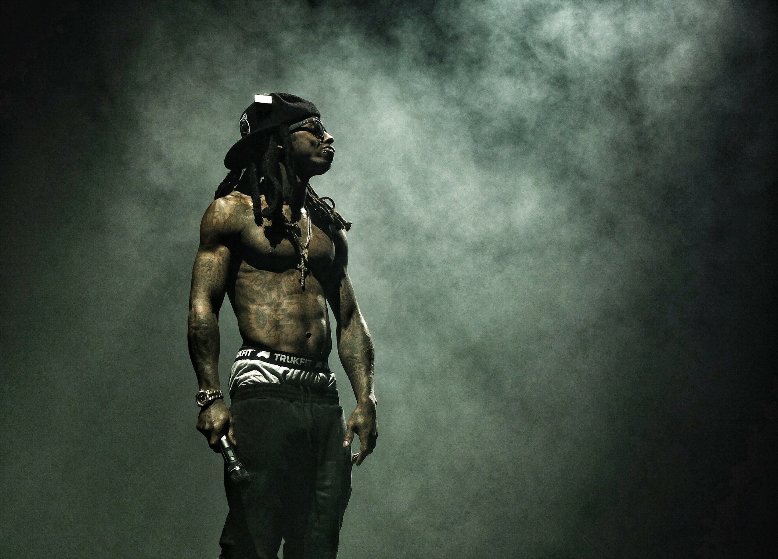 A shirtless man with dreadlocks and sunglasses stands in a smoky environment, looking to the side.