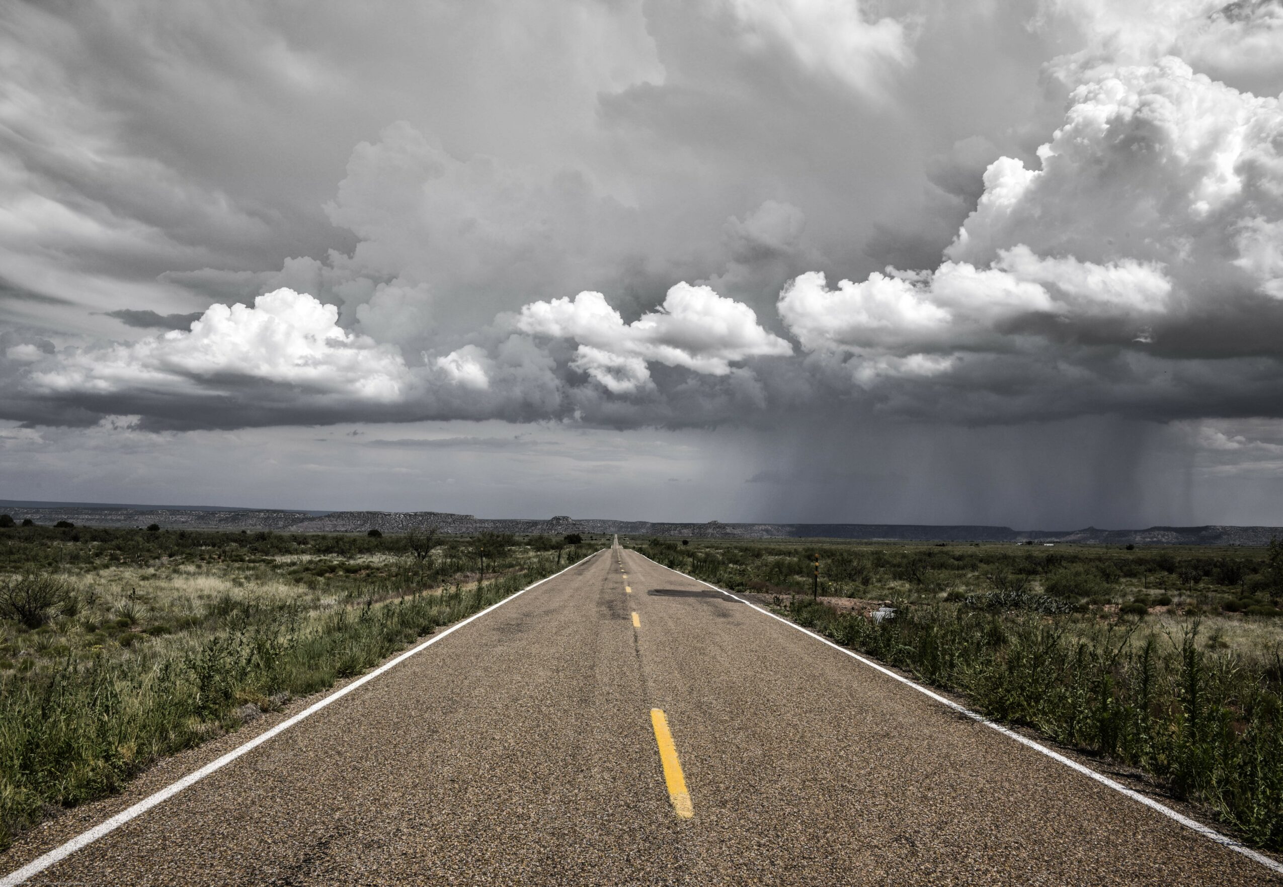 Long empty road stretching into the horizon under a cloudy sky with rain in the distance.