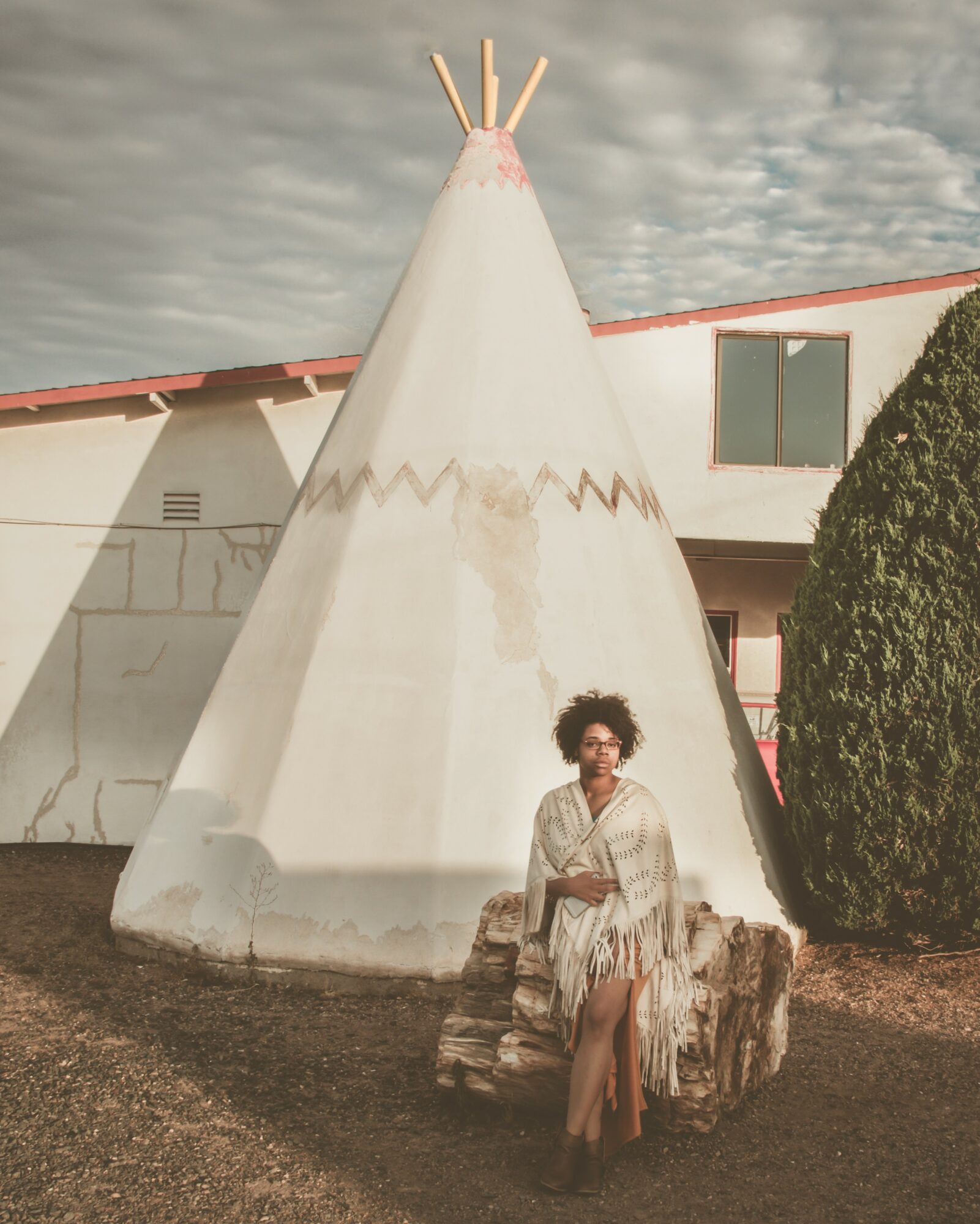 Person sitting on a log in front of a teepee with a building and sky in background.