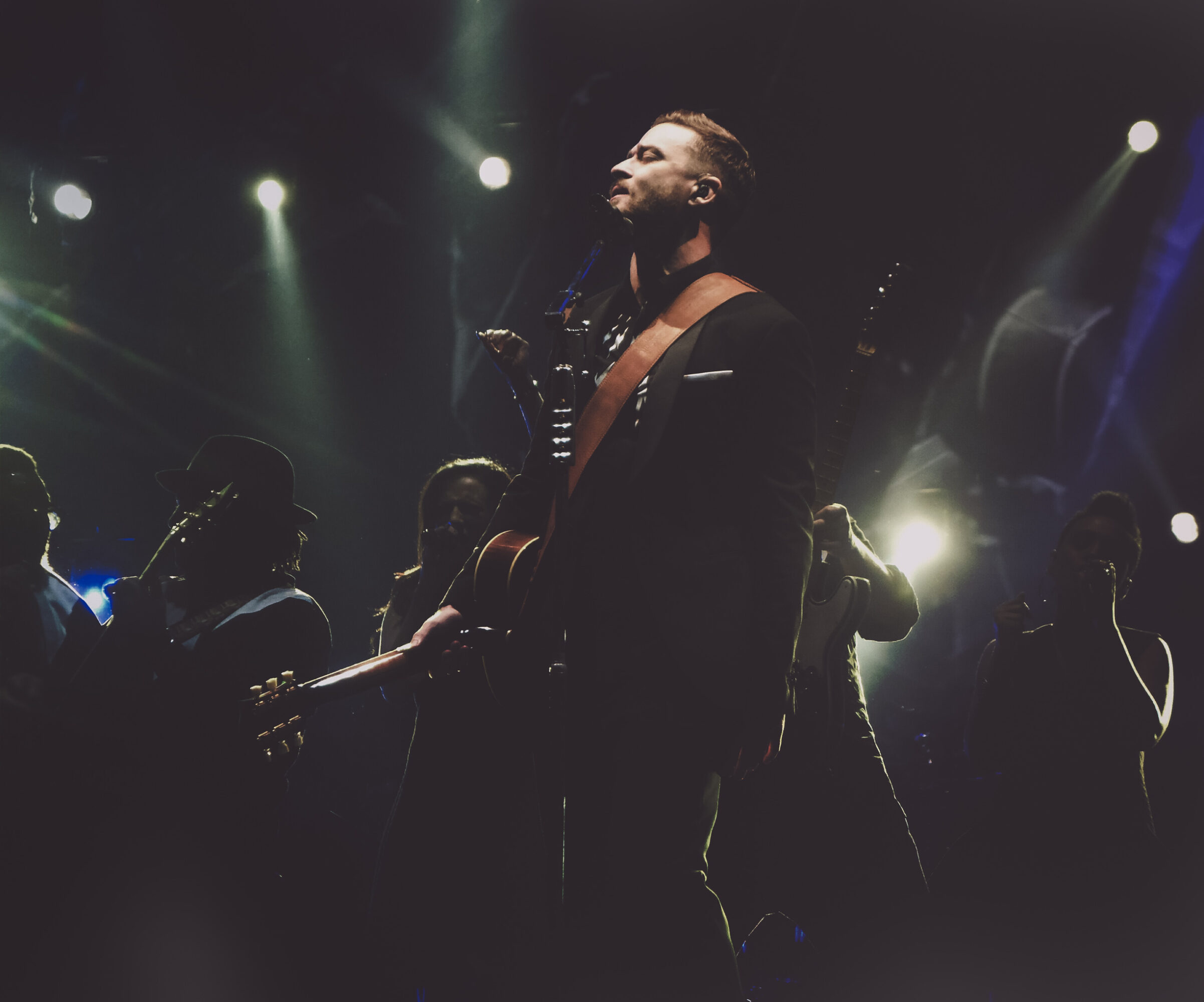 Man with beard and long hair on stage, surrounded by musicians, illuminated by stage lights, dark background