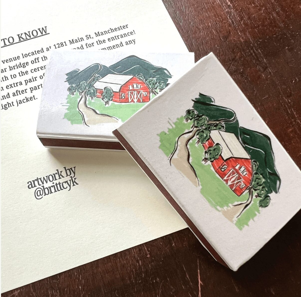 Two matchboxes with illustrations of a red barn, trees, and mountains, placed on a wooden surface with a paper nearby.