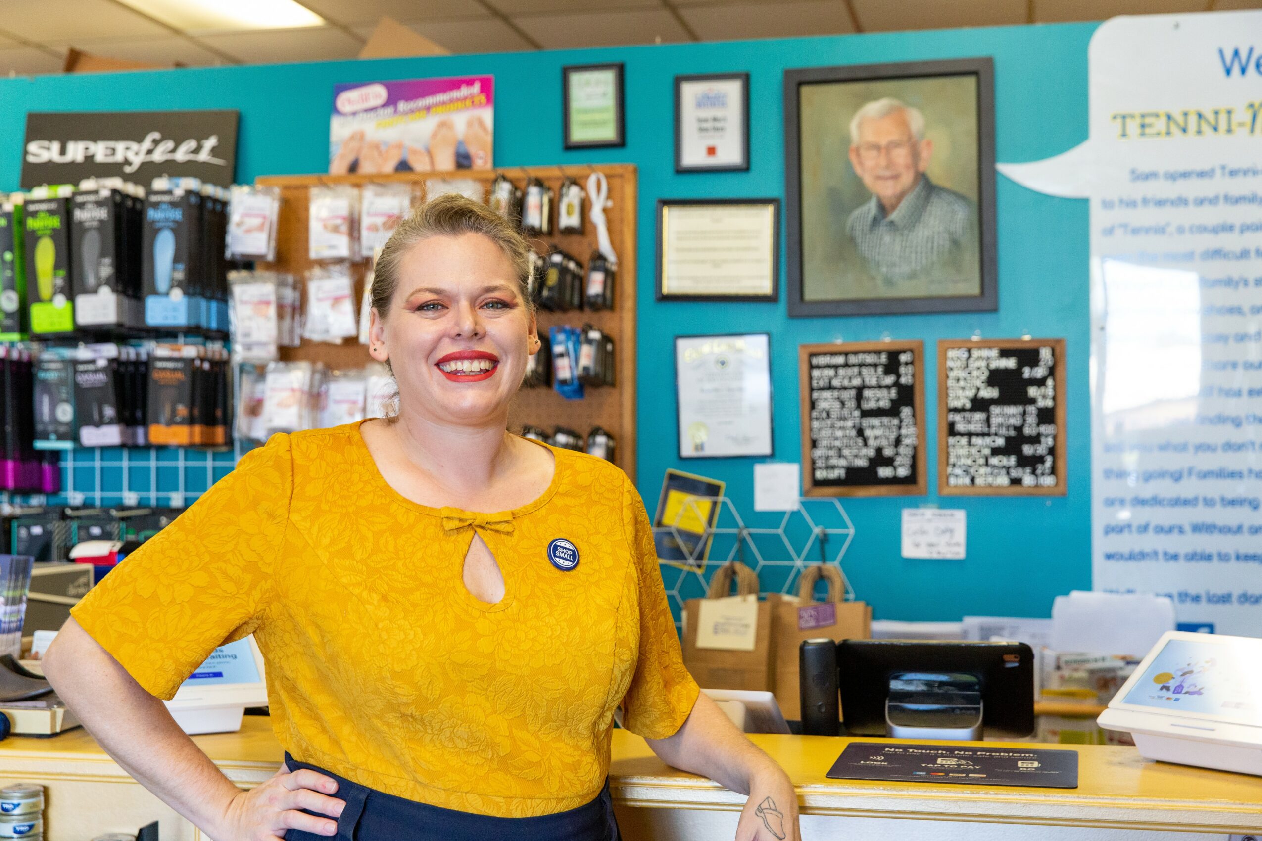 Smiling woman in yellow top standing in a store with framed pictures and products on the wall behind her.