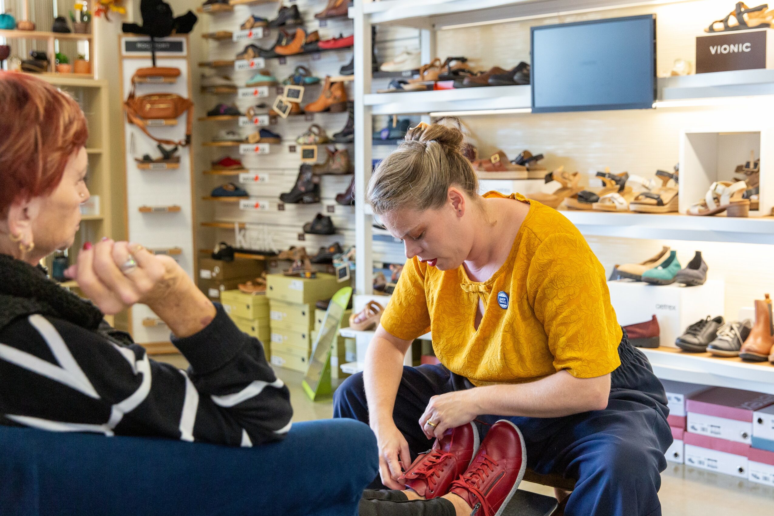 A young woman tries on a red boot while a woman with red hair observes in a shoe store.