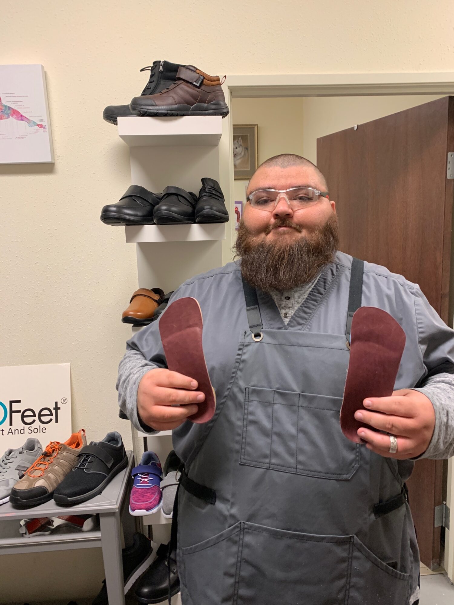 Man with glasses and beard holding a shoe insert in each hand, standing in a shoe store with shelves of shoes behind him.