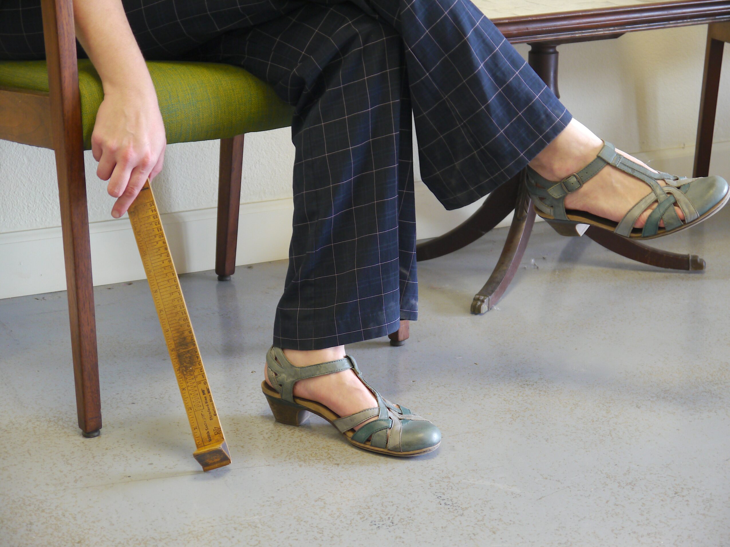 Person sitting on a green chair, holding a measuring tape, wearing plaid pants and green sandals, with a wooden table nearby.