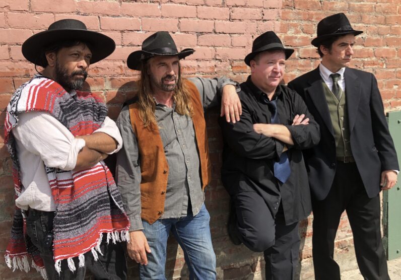 Four men stand against a brick wall, dressed in vintage-style clothing and hats, posing with arms crossed or relaxed.