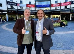 Two men in suits standing outdoors, smiling, giving thumbs-up, in front of a building with digital screens and signage.