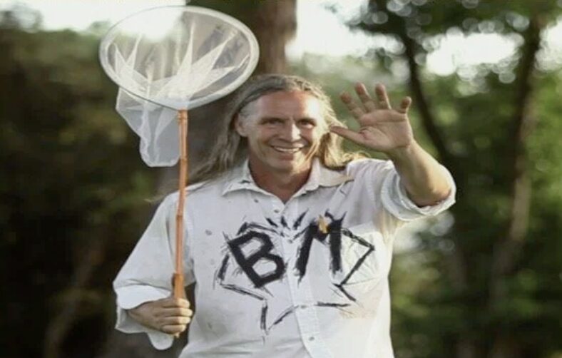 Person smiling and waving outdoors, holding a butterfly net, with trees in the background.