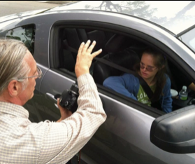 Older man waving at young woman in car window, holding a camera, outdoors on cloudy day.