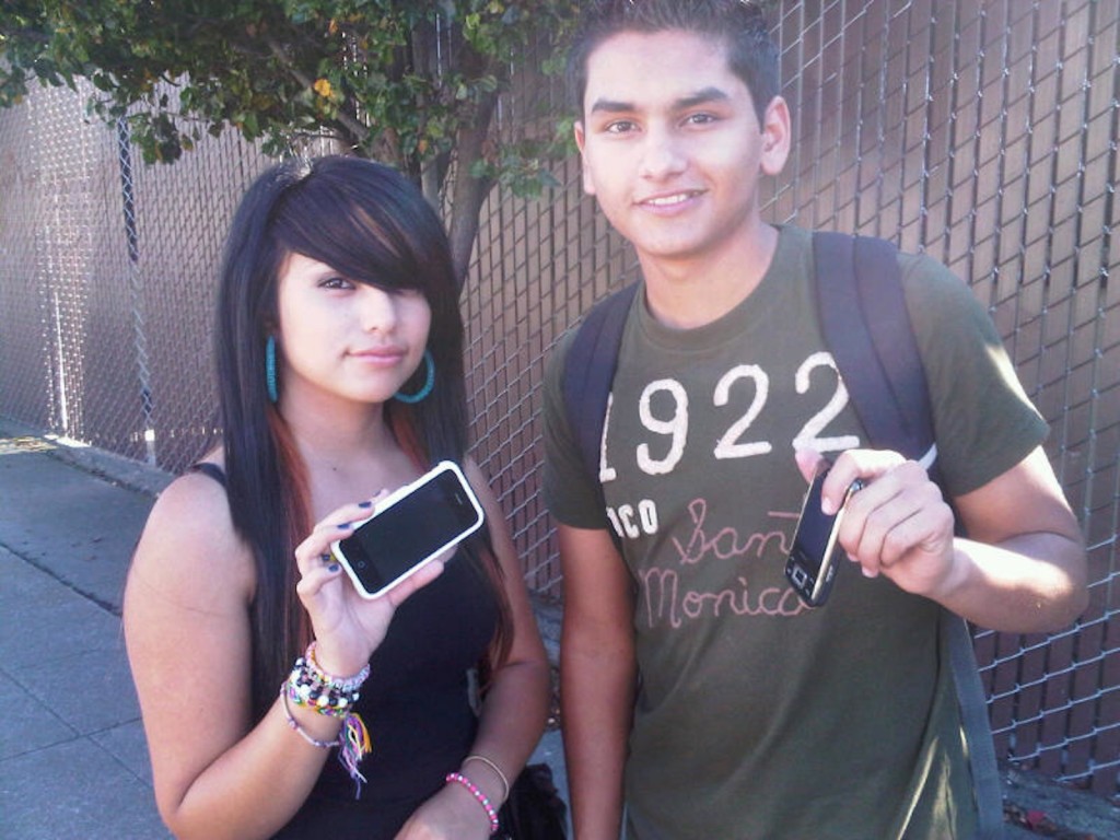 A boy and girl wearing backpacks hold out their cellphones for the camera to capture. 