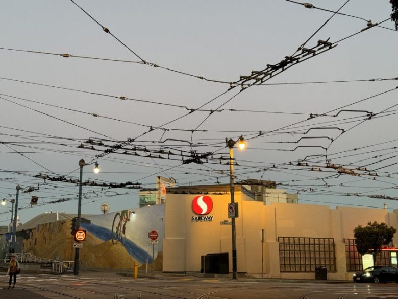 Safeway store at dusk with overhead trolley wires, street signs, and a mural on a nearby wall.